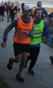A group of runners is seen on a gravel pathway by a scenic waterside. At the forefront, is guide runner Russ wearing an orange high-visibility vest labelled "GUIDE RUNNER" runs alongside Matt in a yellow vest marked "BLIND RUNNER." Behind them, other runners are visible in casual athletic clothing, and the background features serene water and under a clear sky.