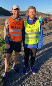 Two runners are standing side by side on a gravel path, smiling for the photo with a backdrop of hills and a calm body of water. The guide runner Russ is on the left wears an orange high-visibility vest labelled "GUIDE RUNNER," while Matt stands on the right sporting a yellow vest marked "BLIND RUNNER." 