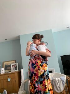 Nia with brown hair tied in a bun and wearing glasses gently cradles two new-born babies in her arms. She is dressed in a vibrant floral dress, holding one baby close to her shoulder while supporting the other in her arms. The setting is a cosy living room with soft blue walls. A wooden cabinet in the background displays framed family photos, congratulatory cards, and decorative heart ornaments. A baby bassinet with a white blanket draped over the side sits nearby.
