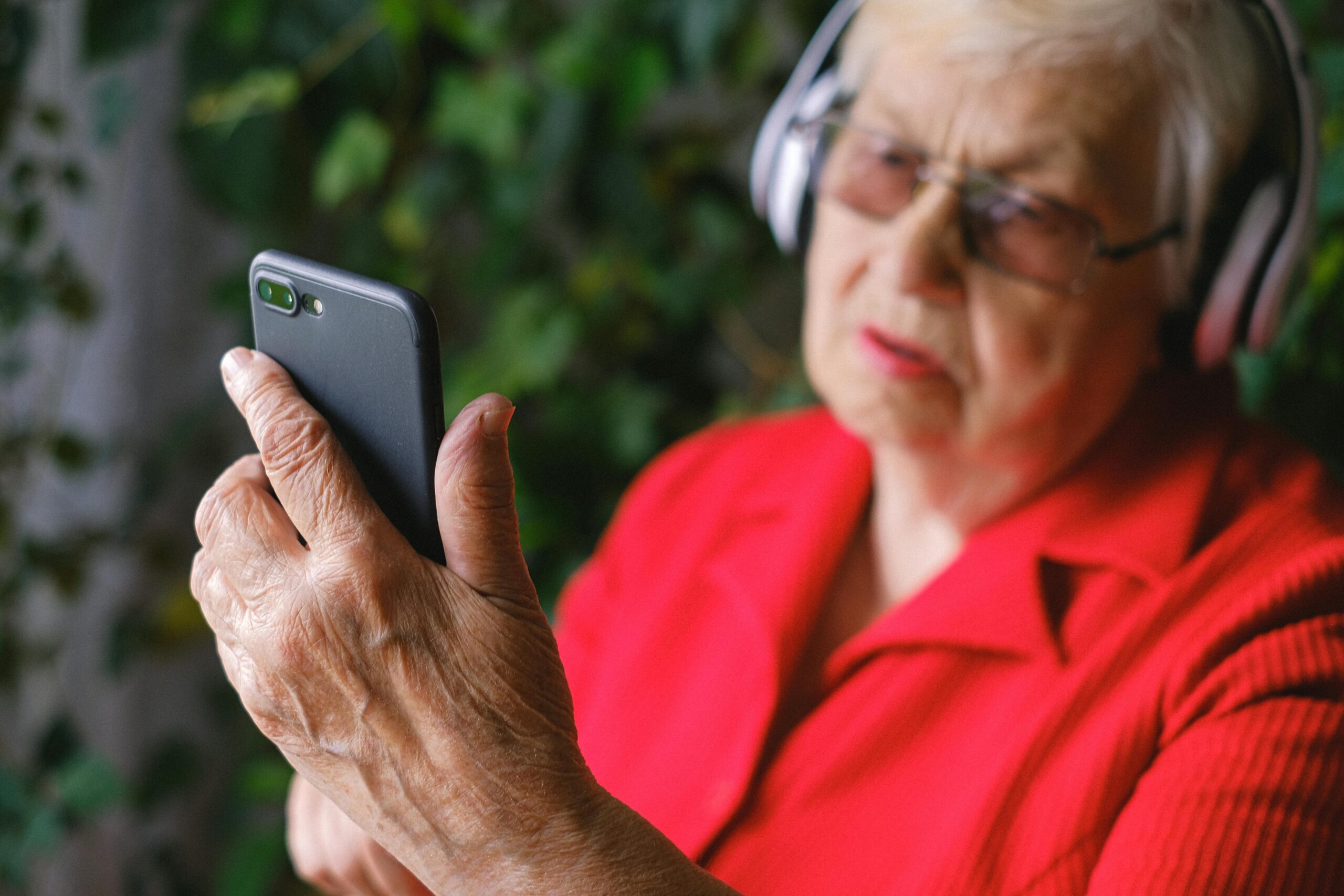 Elderly Woman wearing red and large headphones looking concerned while looking at her mobile phone