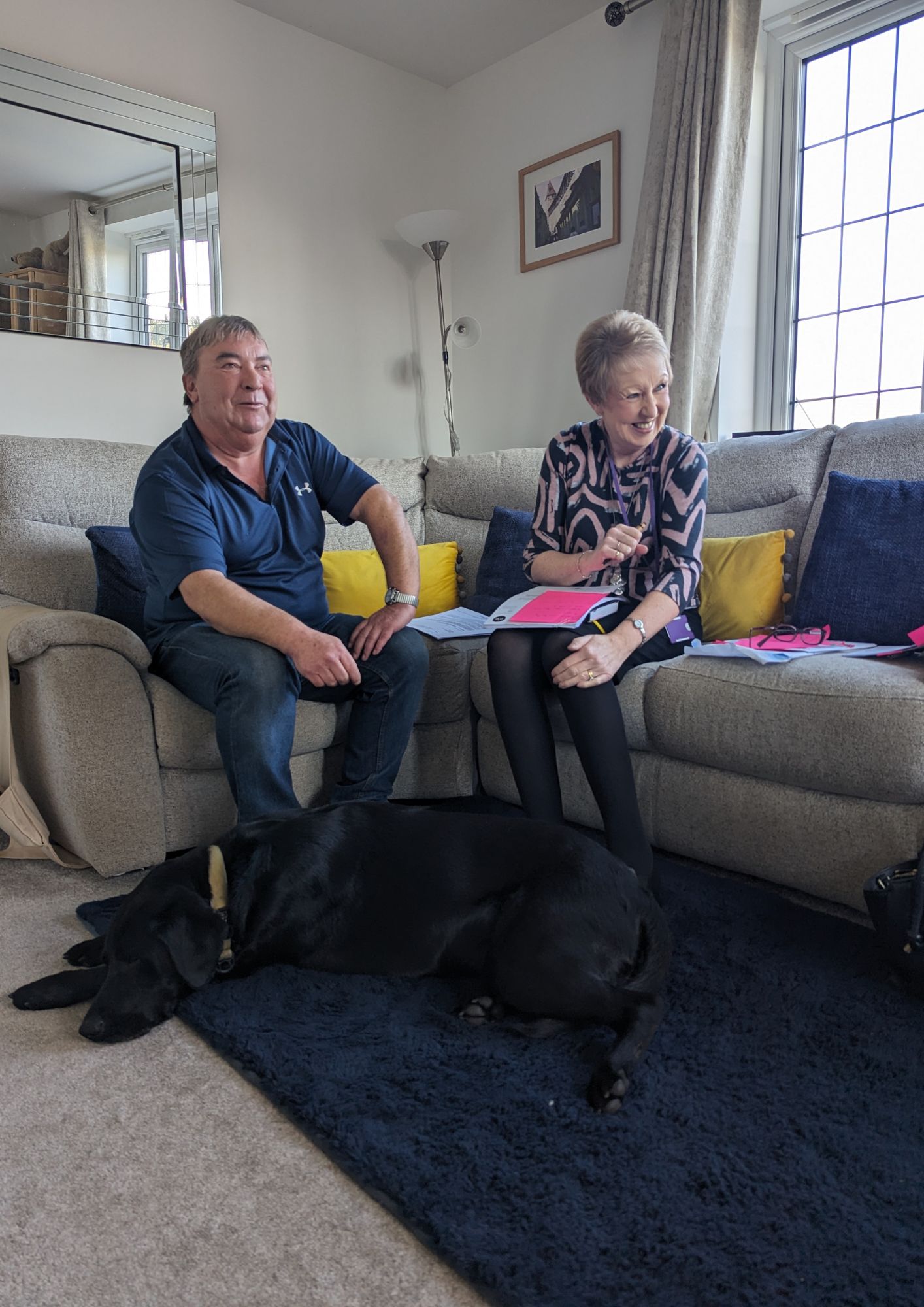 Two people sitting on a sofa during a home visit looking very happy with a black dog laid down on a dark blue rug infront of them