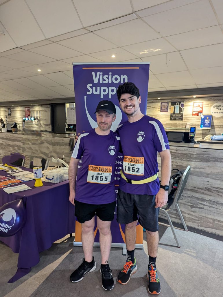 Matt and Lewis stood infront of a Vision Support Banner with their numbers from the 2025 Chester 10K Run visible on their shirts.