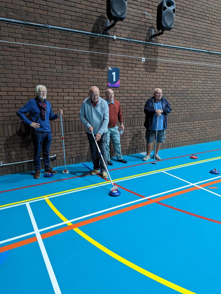Group of older men stood at one end of a curling track looking like they are about to play. They are stood within a large sports hall