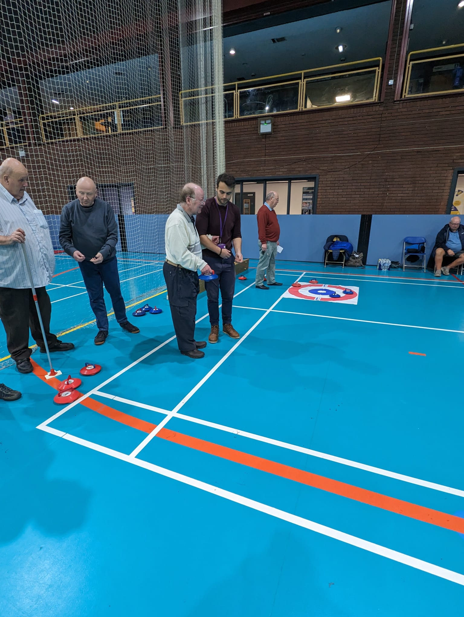 Group of older men stood at one end of a curling track getting instructions on how to play from a younger man. They are stood within a large sports hall that has a balcony area looking over the main hall