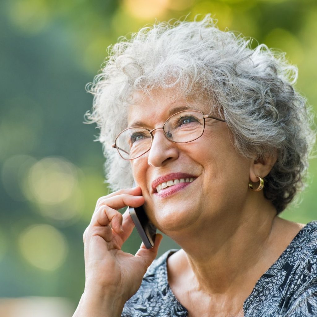 Image of an older woman speaking on her mobile phone. She has grey hair and glasses on with a gold earring in the shape of a ring. She appears to be outside with green foliage visible in the background of the image