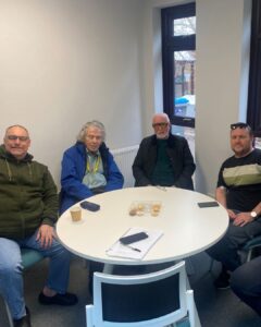 Four men seated around a round white table in a meeting room. Papers, a pen, a mobile phone, a cup, and a tray of biscuits sit on the table. A window and radiator are visible behind them.