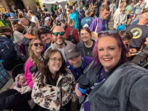 A cheerful group selfie taken at a busy outdoor charity event. In the foreground, a group of around seven adults—men and women stand close together smiling at the camera. One person in front has bright purple hair and is holding the camera, while others lean in.