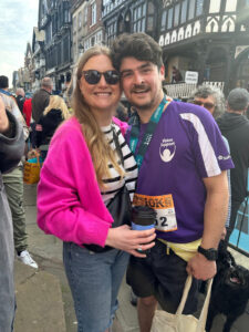 Kate and Lewis stand close together outdoors after a race. Lewis is wearing a purple Vision Support t-shirt, a race medal, and a running bib for a 10K event. Kate wearing sunglasses, a bright pink cardigan over a striped top, and holds a takeaway coffee cup.