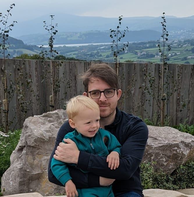 Mat sits outdoors holding his young son in his arms. They are positioned in front of large natural stone rocks and a wooden fence, with a scenic countryside landscape stretching into the distance behind them. Rolling green fields, hills, and a body of water can be seen under an overcast sky. His son smiles while looking slightly to one side, and Mat looks towards the camera