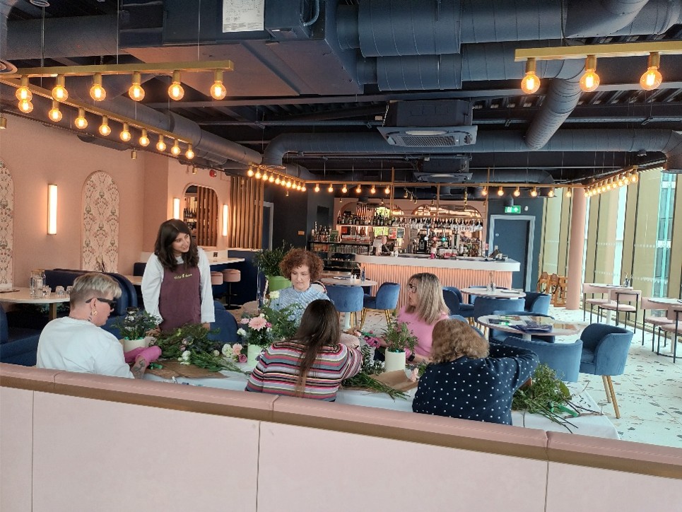 A group of six people take part in a flower arranging workshop around a long table in a modern café. An instructor stands beside the table, offering guidance while participants create floral arrangements using fresh flowers, greenery and pink hat boxes. The bright café features soft pink walls, blue upholstered chairs, round tables and warm hanging lights. Flowers, foliage, wrapping paper and gardening tools are spread across the table as everyone works together on their arrangements.