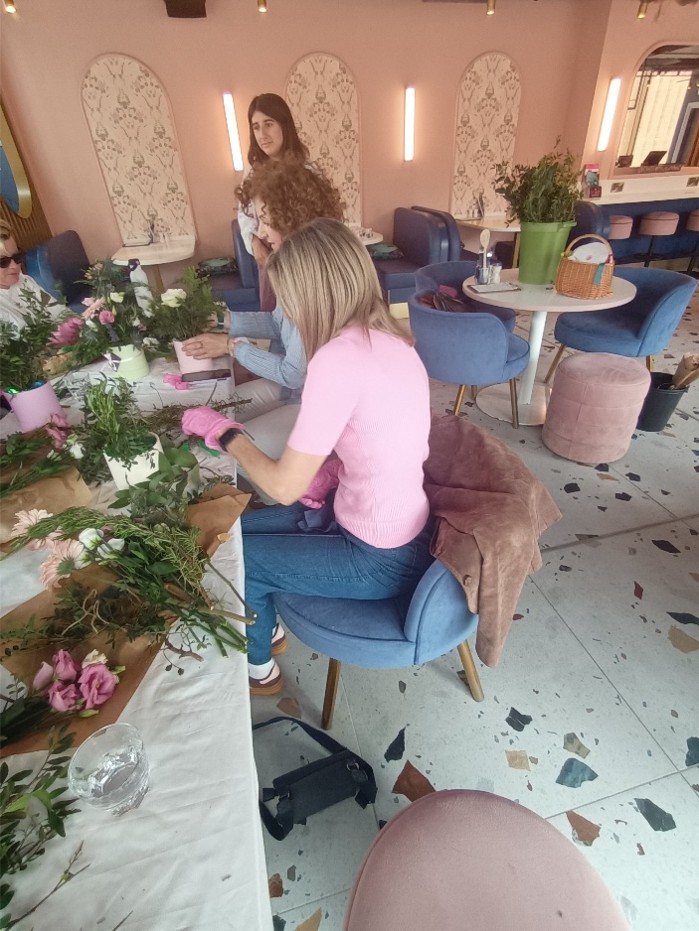 Several people are seated around a long table taking part in a flower arranging workshop inside a stylish café. The table is covered with fresh flowers, greenery, stems, paper wrapping and small plant pots as participants create floral arrangements. In the foreground, a person wearing a pink top and gardening gloves arranges foliage in a white pot. Other participants chat and work together while an instructor stands nearby. The café features soft pink walls, blue upholstered chairs, round tables
